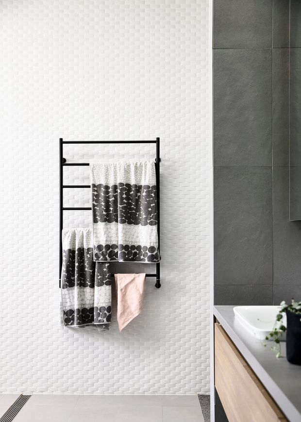 Contemporary bathroom interior featuring a black wall-mounted towel ladder with patterned towels, a white textured mosaic tile wall, large dark grey wall tiles, and a timber vanity with a white basin.