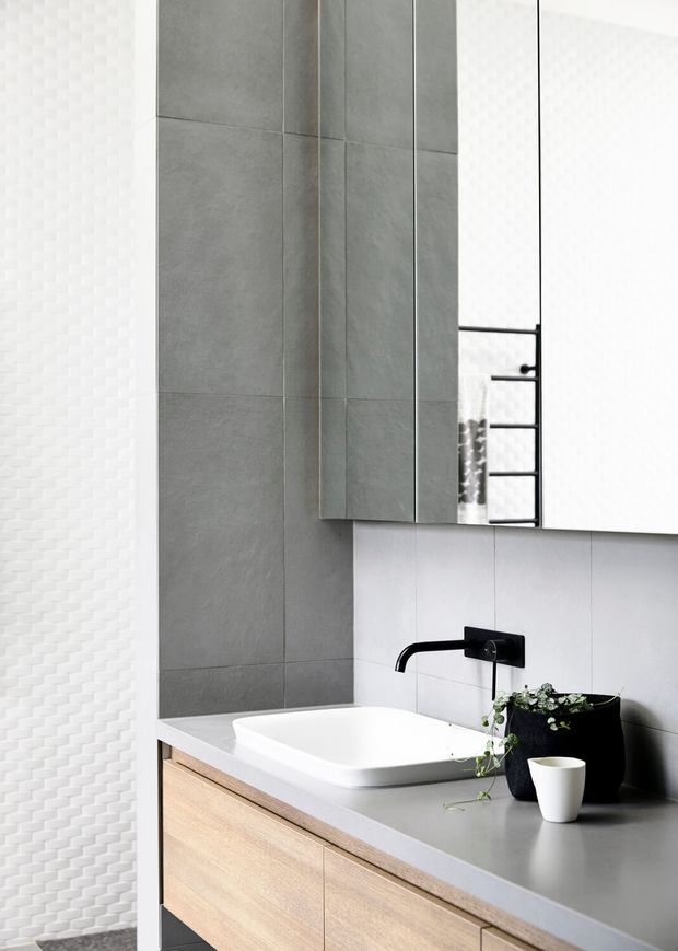 A contemporary bathroom vanity with a white vessel basin set into a grey benchtop, light timber cabinetry, matte black wall-mounted tapware, a large mirrored cabinet, and grey tiled wall surfaces. A white textured feature wall and a small potted plant add subtle contrast.