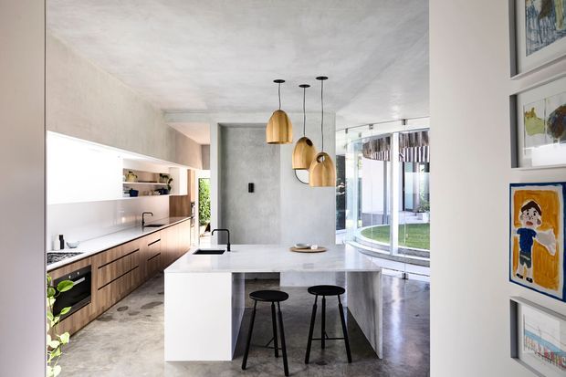Contemporary open-plan kitchen with a large white island, integrated sink and black bar stools. The room features warm timber cabinetry, a pale stone or concrete-look benchtop, textured plaster walls and ceiling, pendant lights with brass-toned shades, polished concrete flooring, and expansive curved glazing overlooking a courtyard or garden. Framed children's artwork is displayed on the adjacent wall.