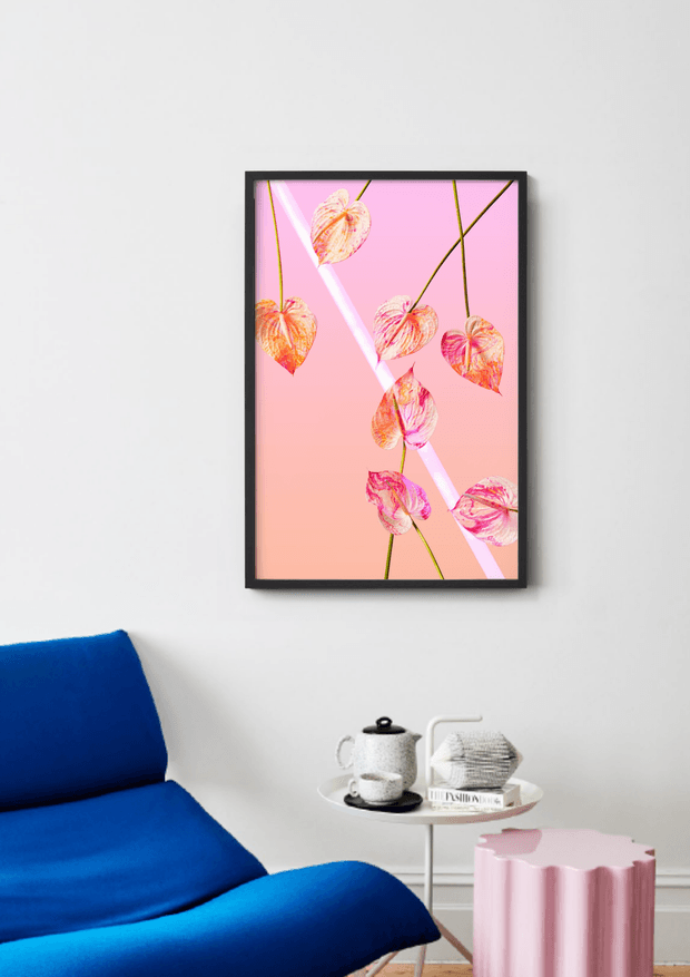 A bright, minimalist sitting area with a vivid cobalt-blue lounge chair, a small round white side table holding a teapot and cup, and a pink scalloped cylindrical stool. A framed botanical artwork with pink and coral leaves hangs on the white wall above the table. The space has a playful, contemporary colour palette and simple furnishings.