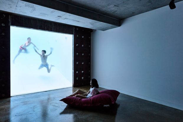 A dark, minimalist media room with a large wall-sized projection screen displaying swimmers, a person reclining on a beanbag, polished concrete flooring, exposed concrete ceiling beams, and a dark climbing-wall panel framing the screen.