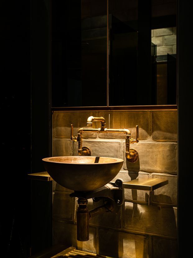 A dark, atmospheric bathroom vignette featuring a freestanding vessel basin with exposed industrial-style copper or brass plumbing, wall-mounted taps, and slim metal side shelves. The basin is set against a textured block-like metallic or stone wall finish, with a large dark reflective mirror above. Warm directional lighting creates a dramatic, luxurious mood.