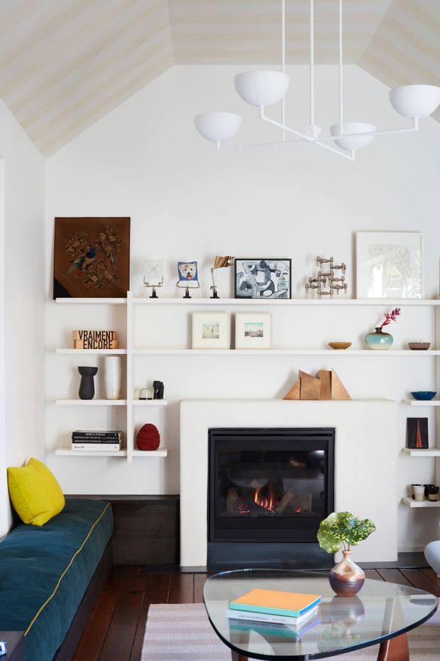 A bright, carefully styled living room centred on a contemporary built-in fireplace and white open shelving. The room has a vaulted ceiling with pale timber lining, white walls and joinery, dark timber floorboards, a deep teal sofa with a bright yellow cushion, a glass coffee table, artworks, ceramics and books. A white sculptural pendant hangs overhead, creating a refined yet inviting interior.