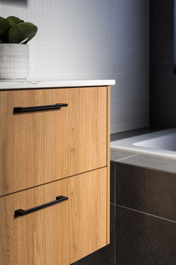 Close-up view of a contemporary bathroom vanity with warm timber-grain cabinetry, slim black bar handles, and a light stone benchtop. A textured dark tiled wall and the edge of a white bathtub are visible beside it, with a potted plant adding a small natural accent.