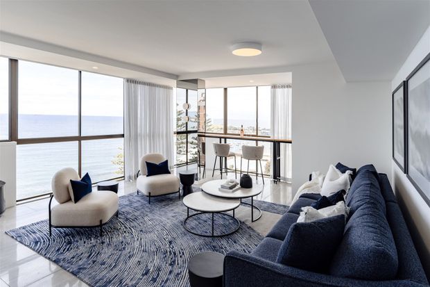 Bright contemporary living room overlooking the water, with expansive floor-to-ceiling glazing, a dark blue sectional sofa, cream lounge chairs, round nesting coffee tables, and a patterned blue rug. A compact kitchen or bar counter with stools is visible behind the seating area. The room has a clean, refined coastal feel and abundant natural light.
