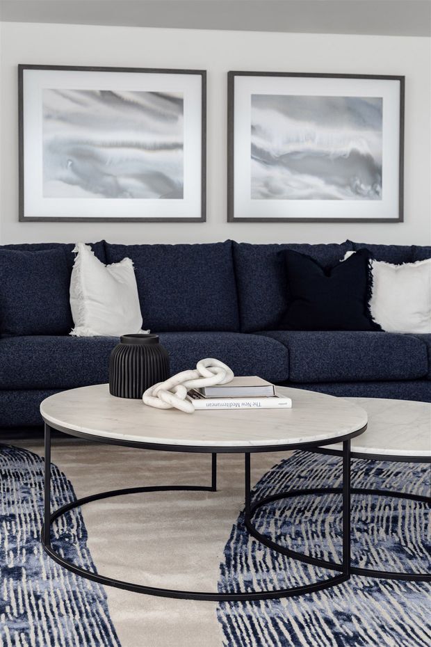 A refined living room vignette featuring a deep navy upholstered sectional sofa with white and dark cushions, two abstract monochromatic artworks in dark frames, and overlapping round coffee tables with pale stone tops and slim black metal bases. Layered blue-and-cream patterned rugs add texture beneath the tables, while a dark ribbed ceramic vase and sculptural white chain ornament sit on the main table.