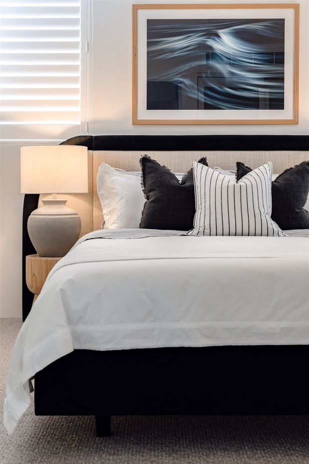 A neatly styled contemporary bedroom featuring a black upholstered bed with a tall headboard, crisp white bedding, black and white decorative pillows, and a light wood bedside table with a ceramic lamp. White horizontal blinds cover the window, while an abstract blue-grey artwork in a natural wood frame hangs above the bed. The palette is monochromatic with warm timber accents and soft, layered textures.