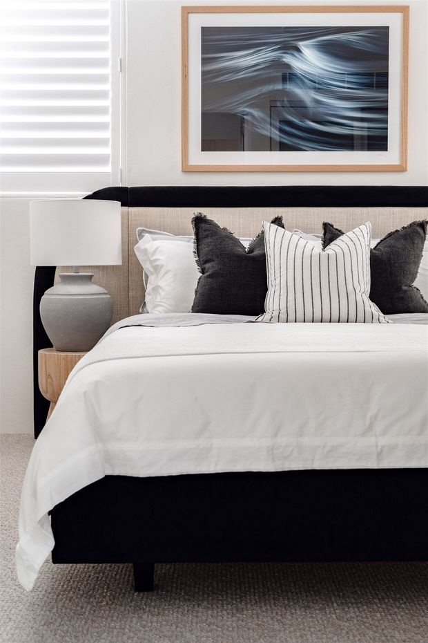 A refined bedroom scene with a neatly made bed, black upholstered bed base and headboard, white bedding, charcoal and striped accent cushions, and a light timber bedside table. A grey ceramic lamp sits beside the bed, while a framed abstract artwork hangs above the neutral upholstered headboard. White horizontal blinds and soft neutral finishes create a calm, contemporary monochromatic interior.