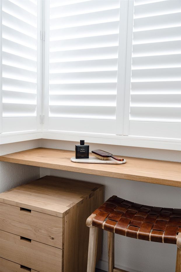 Bright, compact built-in window seat or dressing nook with white plantation shutters, a light timber corner shelf and bench, matching timber drawer cabinetry, and a small woven leather stool. The space has a clean, warm, minimalist fit-out with abundant filtered daylight.
