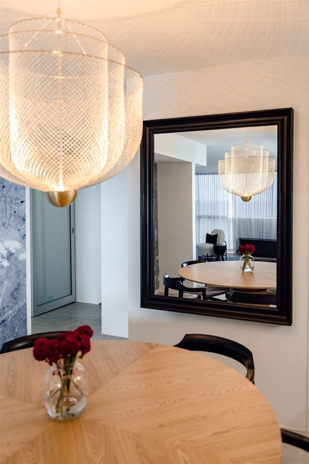A refined dining area with a round light-wood dining table and black chairs, prominently featuring a large woven pendant light and a substantial dark-framed wall mirror. The mirror reflects additional seating, sheer curtains, and the pendant light. Pale walls, a grey stone-like vertical surface, and a cool grey tiled floor create a contemporary, softly lit interior.