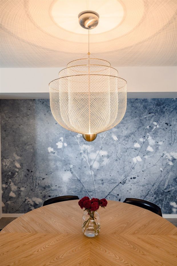 A refined dining area centered on a large round light-wood dining table with a geometric parquet-style grain pattern, dark curved dining chairs, and a small vase of red flowers. A sculptural tiered mesh pendant light hangs above, with a dramatic blue-grey veined stone or marble-look feature wall behind it and a pale ceiling.