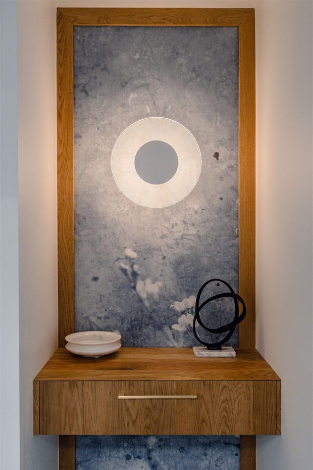 A narrow built-in timber display and storage cabinet set against a mottled blue-grey feature panel. The warm wood surround and drawer feature a prominent grain, with a circular wall detail above, a shallow ceramic bowl, and a black abstract sculpture arranged on the countertop. The composition is minimalist and gallery-like.