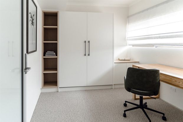 Bright, compact home office with a built-in white storage cupboard, open timber shelving, a long timber desk beneath a large window, and a black swivel office chair. The room has light carpet, white walls, clean modern detailing, and abundant natural light.