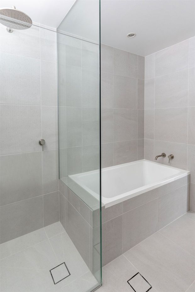 Bright contemporary bathroom featuring a frameless glass shower screen with overhead rainfall shower, a built-in white bathtub, pale grey tiled walls and floor, chrome fixtures, and recessed ceiling lighting.