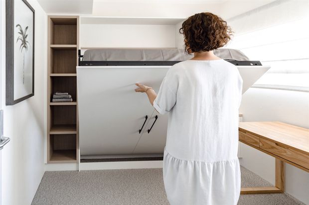 Bright compact multifunctional room featuring a wall-mounted fold-down bed or Murphy bed in an open position, integrated dark timber shelving, a built-in timber desk beneath a large window, light carpet flooring, and white walls. A person stands in front of the bed mechanism.