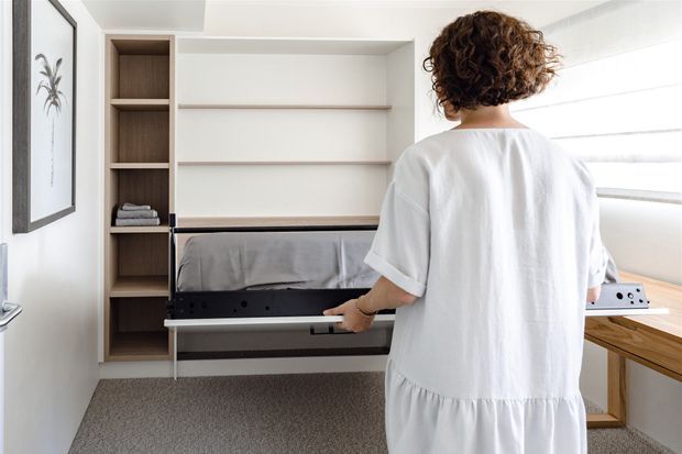Bright compact bedroom with a wall-integrated fold-down bed being operated by a person. The room includes built-in open shelving, simple horizontal display shelves, light neutral walls, grey carpet, and a timber-framed window seat or desk beside broad horizontal glazing. The design is functional, minimalist, and space-efficient.