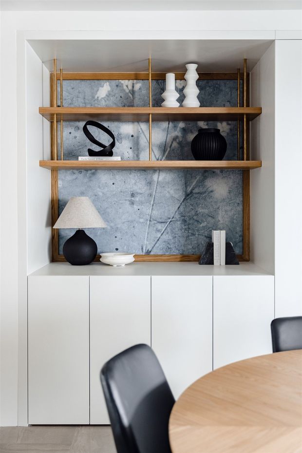 A refined built-in dining room storage and display unit with white cabinetry, warm timber shelving, slim brass supports, and a textured grey-blue feature backing. The shelves display monochrome ceramics, sculptural objects, books, and a black table lamp. A round timber dining table and black upholstered chairs occupy the foreground. The overall composition is clean, contemporary, and restrained with a sophisticated neutral palette.