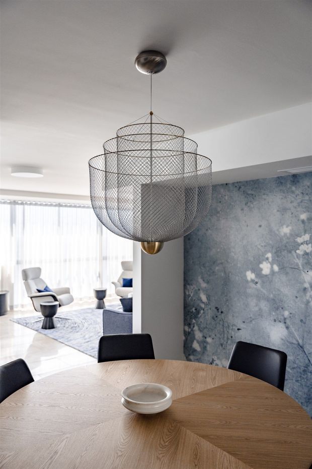 Contemporary dining area featuring a large round light-wood dining table with black upholstered chairs and a sculptural tiered wire-mesh pendant light. The space opens into a bright living area with lounge chairs and sheer curtains, while a dramatic blue-grey textured feature wall adds contrast.