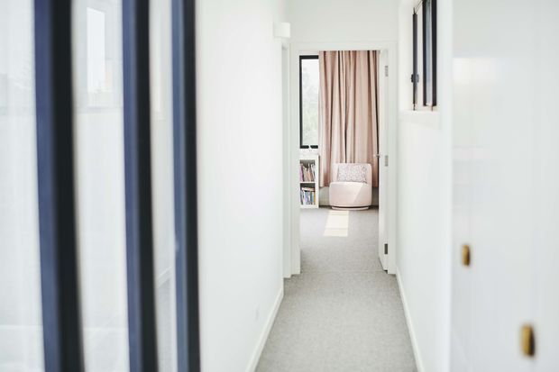 A bright, narrow residential hallway with white walls, pale carpet, dark-framed glazing or doors along the left, and a small window on the right. At the end, the corridor opens toward a softly furnished sitting or bedroom area with blush curtains, a pale armchair, and a small bookcase.