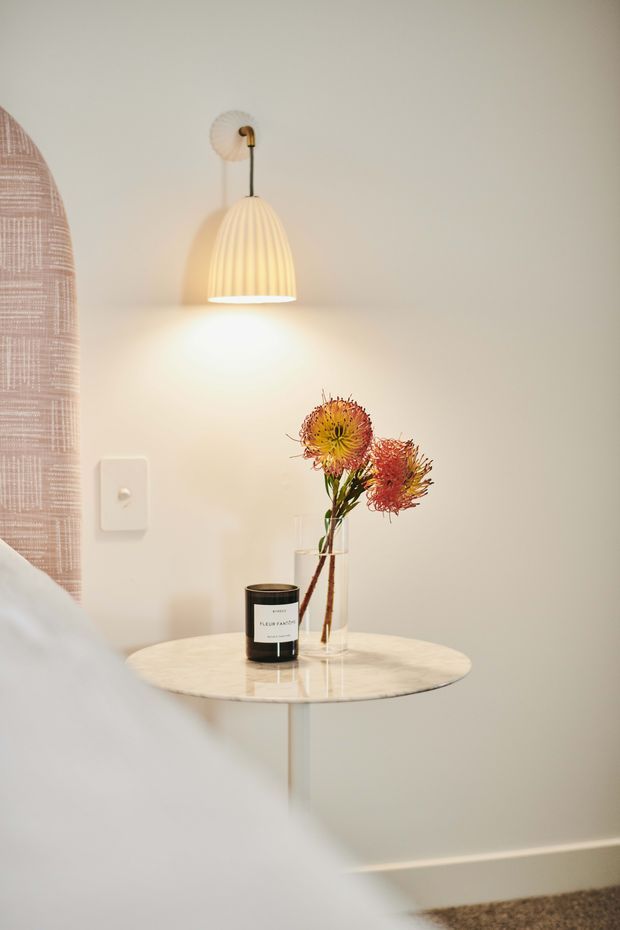 A softly styled bedroom corner featuring a small round marble-topped side table with a slender pedestal base, a glass vase of dried flowers, and a candle. Above it is a warm ribbed pendant wall light with a brass-toned fitting. The pale neutral wall, upholstered curved bed or headboard edge, and muted carpet create a calm, refined atmosphere.