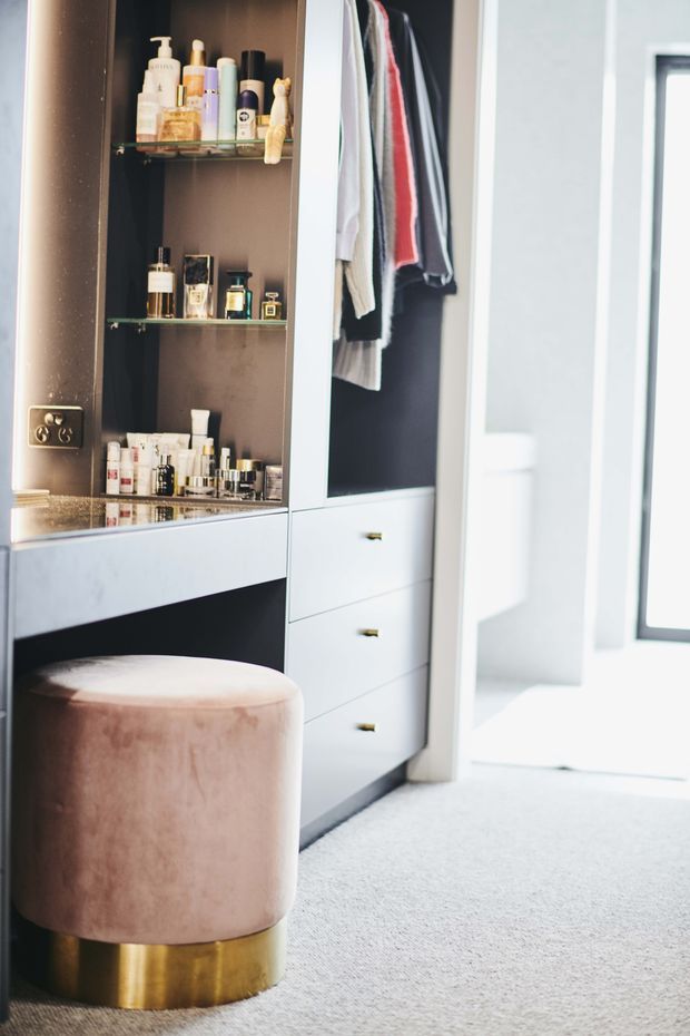 Bright dressing room or walk-in wardrobe featuring built-in white drawers, an open hanging section, and a dark mirrored display cabinet with glass shelves for cosmetics and accessories. A round blush velvet stool with a brass base sits on light carpet, with a bright adjoining space visible through the doorway.