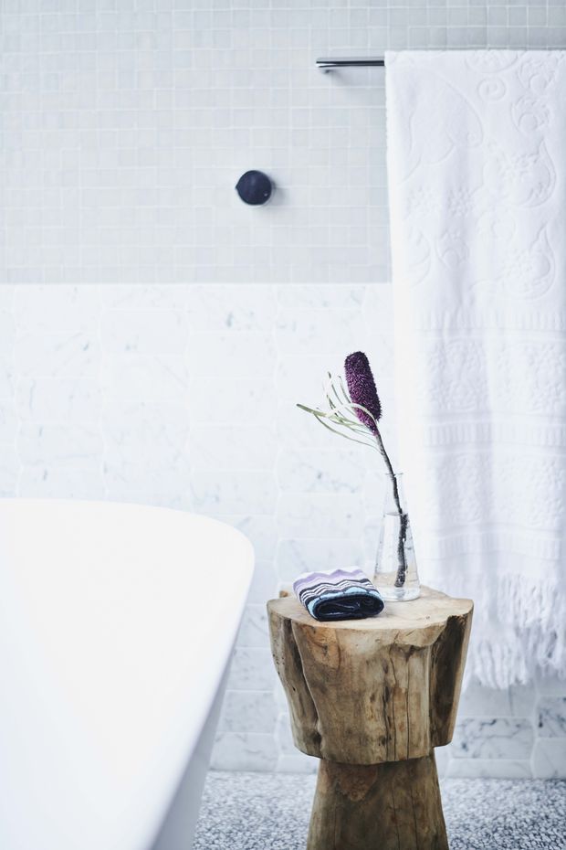 A bright, serene bathroom featuring a freestanding white bathtub, pale white tiled walls, a white marble-look tiled lower wall, dark towel rail and fixture, and a rustic carved timber stump side table holding folded towels and a glass vase. Small grey mosaic tiles cover the floor, with a white textured towel hanging at right.