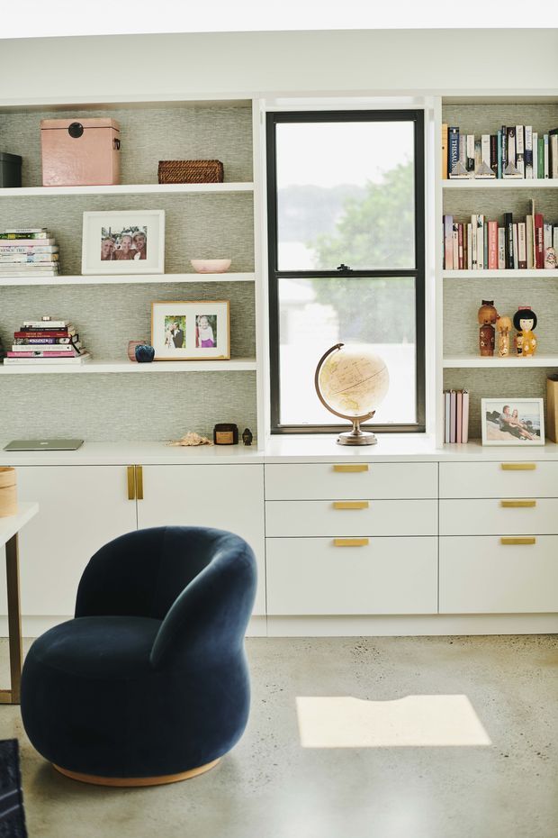 A bright, carefully styled home office or study with a full wall of built-in open shelving and pale cabinetry surrounding a central black-framed window. The shelves display books, framed photographs, baskets and small decorative objects, while the lower joinery provides drawers and cupboards with brushed brass pulls. A deep navy upholstered lounge chair sits in the foreground on a light polished concrete or terrazzo-like floor. The overall look is refined, warm and contemporary with mid-century-inspired furnishings.