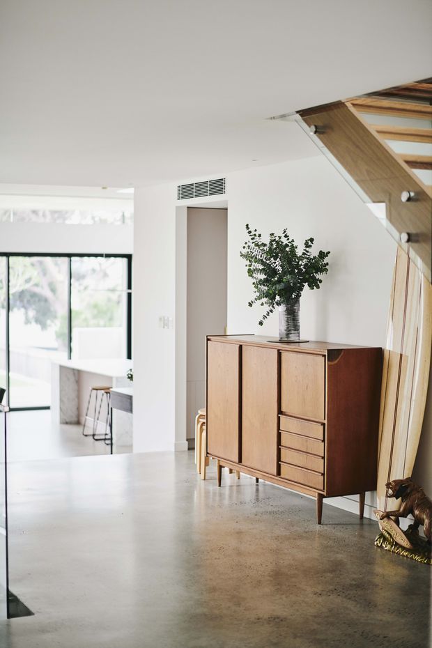 A bright, minimalist interior hallway or entry area with a polished concrete floor, white walls, black-framed glazing, and a mid-century timber sideboard on tapered legs. A timber-framed stairway with open or glazed balustrade occupies the right side, while a kitchen or dining area is visible in the background. A leafy branch arrangement and small decorative object sit on the sideboard.