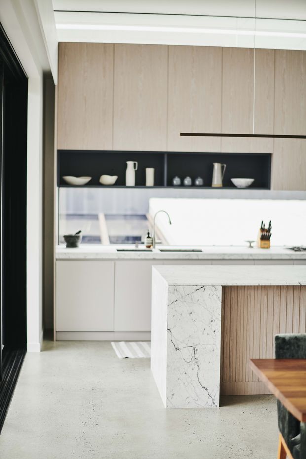 Contemporary kitchen with pale timber overhead cabinetry, white lower cabinets, a dark open display niche, and a large marble-clad island with vertical timber detailing. The space has a polished concrete floor, minimalist pendant lighting, and a bright, refined neutral palette.