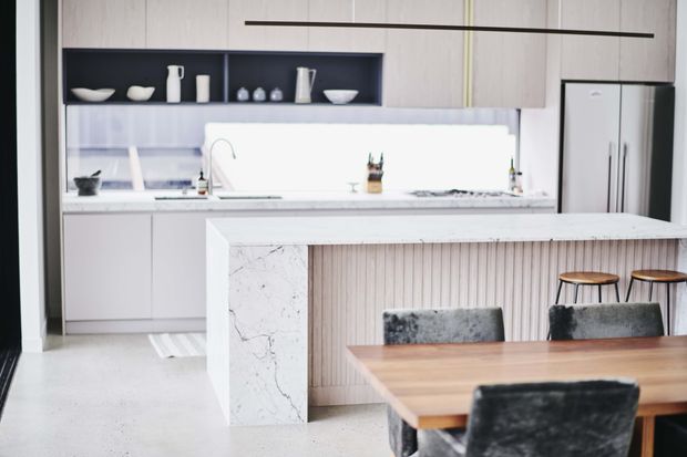 Bright contemporary open-plan kitchen and dining area with a large white stone island, integrated dining table, pale timber vertical wall panelling, white cabinetry, dark open shelving, and a stainless-steel refrigerator. Soft grey upholstered dining chairs and timber stools add contrast.