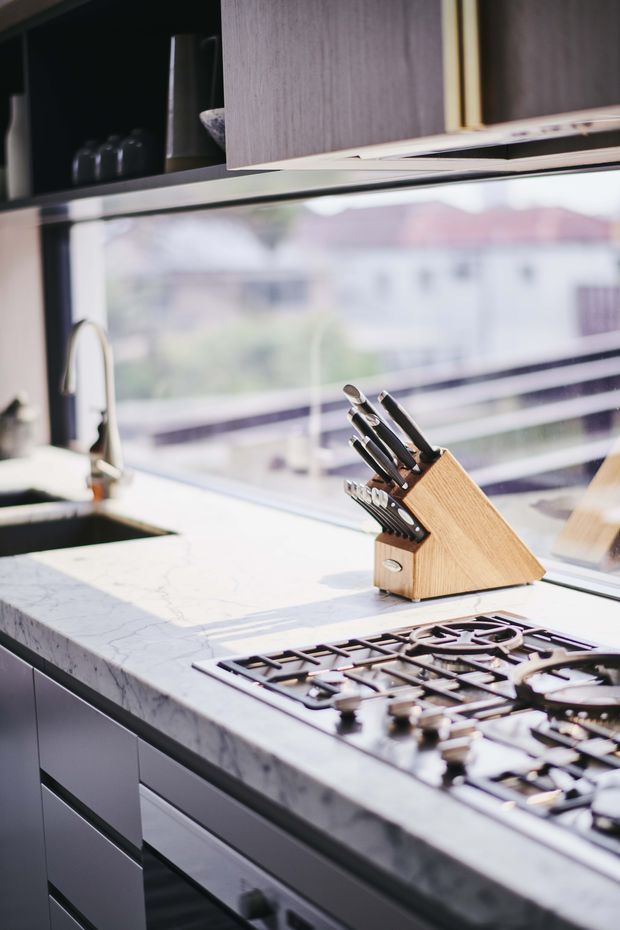 Close-up view of a contemporary kitchen featuring a pale veined stone benchtop, integrated black gas cooktop, undermount sink with tall curved tap, dark cabinetry, open shelving, and a timber knife block. Large windows provide bright natural light and an urban outlook.