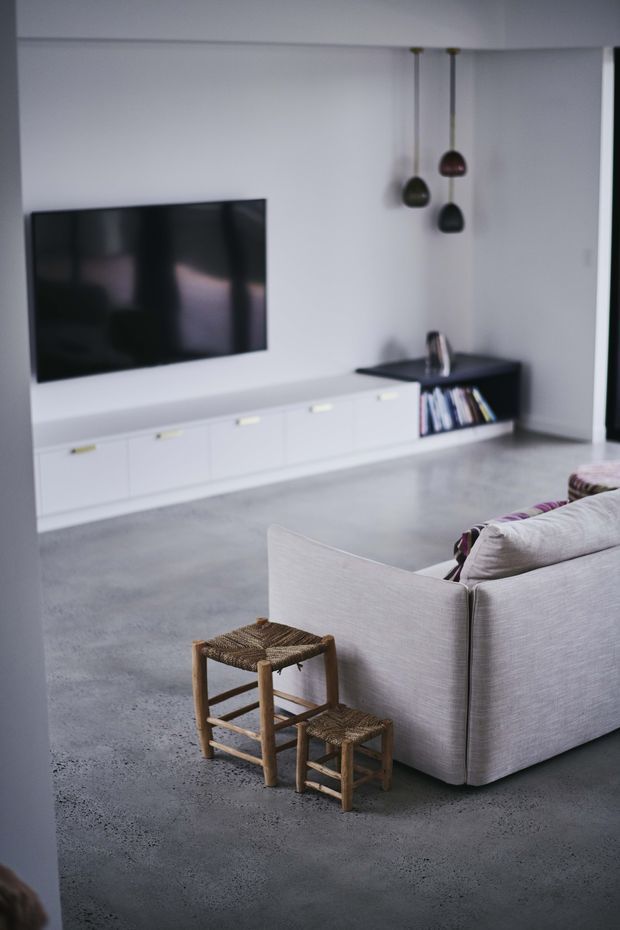 Contemporary living room with a minimalist white built-in media console, wall-mounted television, polished grey concrete floor, and a light upholstered sectional sofa. Three small pendant lights hang above a dark built-in shelf containing books, while rustic woven stools add natural texture in the foreground.