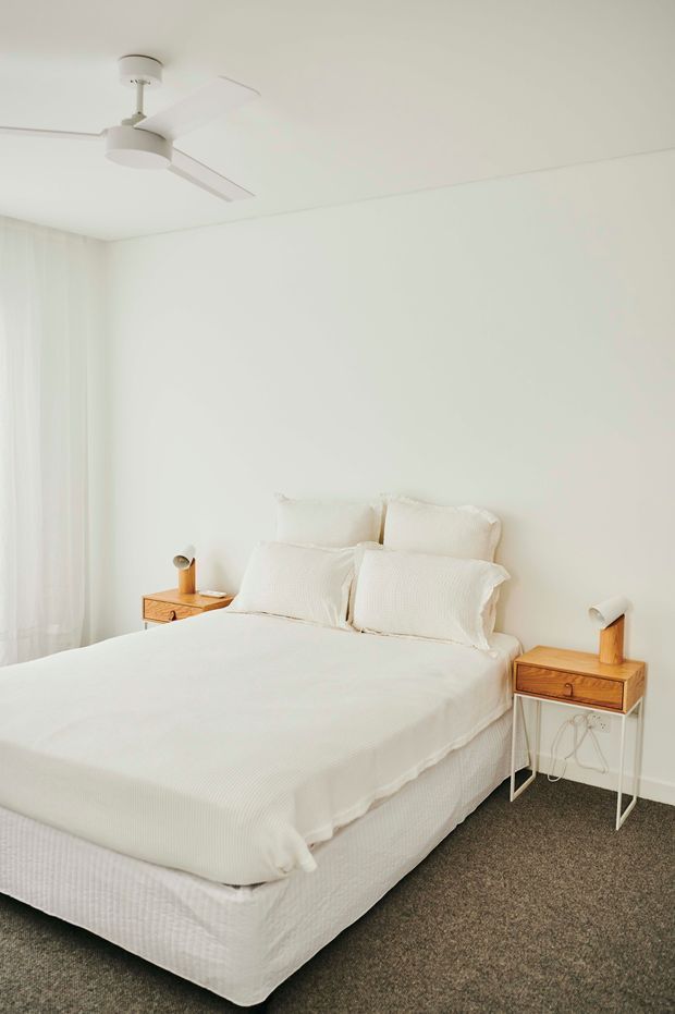 A bright, minimalist bedroom with a white upholstered bed dressed in white bedding, matching wooden bedside tables, and simple integrated lamps. Full-height sheer curtains soften the light, while a white ceiling fan and neutral carpet complete the calm, uncluttered interior.