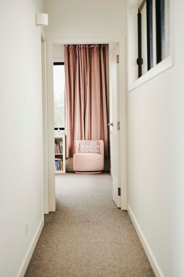 A bright residential hallway with white painted walls, white trim and doors, and beige carpet leading into a small sitting or bedroom area. The adjoining room contains a blush-pink upholstered armchair, patterned cushion, full-length dusty-rose curtains, and a small bookshelf. A framed wall mirror or artwork is partially visible on the right.