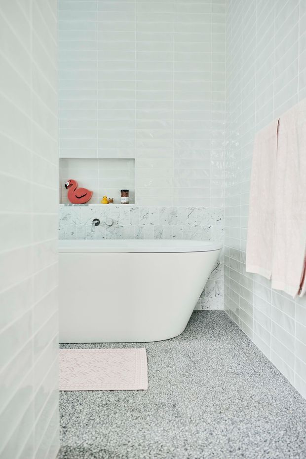 Bright, compact bathroom featuring a freestanding white bathtub, pale glossy wall tiles, a marble-look tiled bath surround, and a terrazzo-style grey floor. A recessed shelf contains small decorative items, while pale towels hang on the right wall.