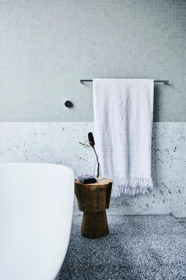 A softly lit contemporary bathroom featuring a white freestanding bathtub, pale green-grey small-format mosaic wall tiles, a lower band of white veined marble-look tiles, and a dark grey terrazzo floor. A white fringed towel hangs from a slim metal rail above a rustic timber stool or side table.