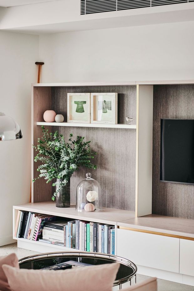 A refined built-in living room media and display wall with dark timber-grain backing, pale cabinetry, open shelving, books, framed artwork, decorative objects, greenery, and a wall-mounted television. A chrome-framed lounge chair and soft furnishings are visible in the foreground. The overall palette is light neutral with warm wood accents and restrained contemporary styling.