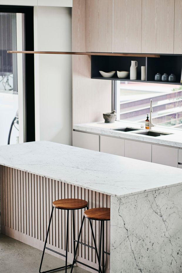 Contemporary kitchen interior featuring a large white marble island with a vertically fluted timber front, two timber-seat black metal bar stools, pale timber cabinetry, a dark open display niche, and a matching marble worktop around the sink. A large window provides an urban outlook and bright natural light.