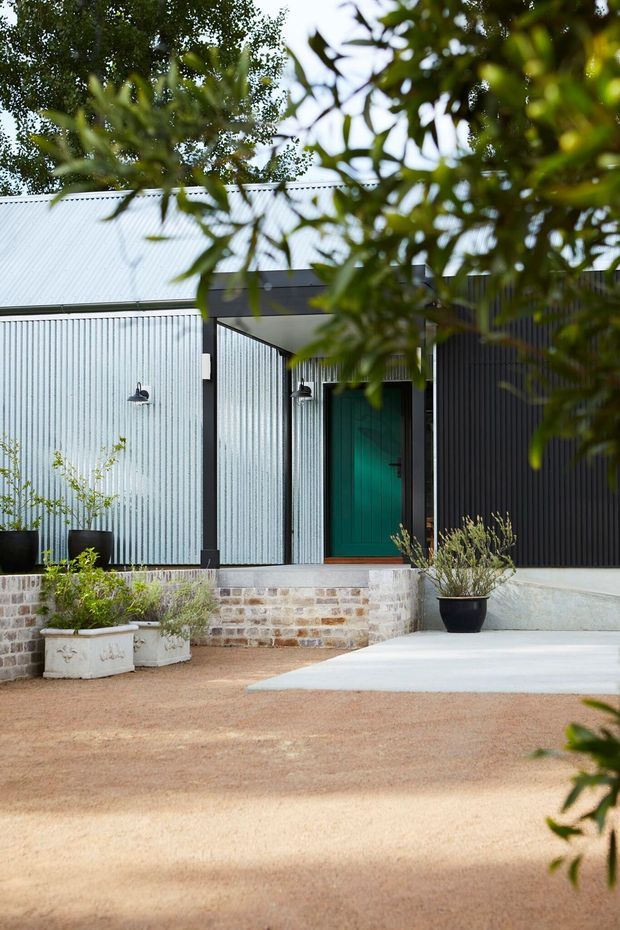 Exterior view of a contemporary single-storey home entrance, framed by leafy trees. The facade combines pale corrugated metal cladding with dark vertical cladding, a covered entry porch, a vivid teal-green front door, exposed brick retaining walls, and simple landscaped planters around a broad gravel forecourt.