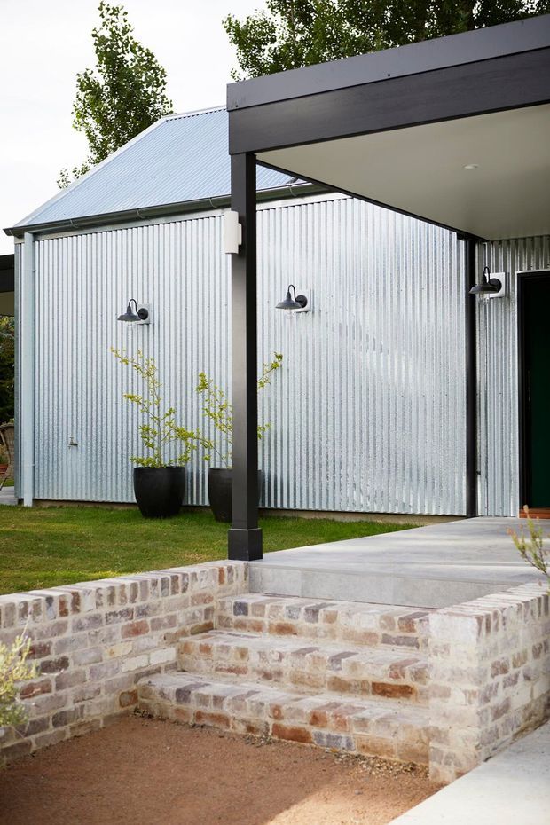 Exterior view of a contemporary residential building with corrugated metal wall cladding and a pitched metal roof. A dark timber or metal-framed covered entry porch leads to a concrete path and steps, bordered by low reclaimed-looking brick masonry walls. Black wall-mounted exterior lights and potted plants add detail to the landscaped approach.