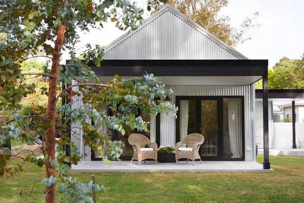 Front exterior view of a compact gabled dwelling with vertically ribbed light-grey metal cladding, a black projecting flat-roofed porch canopy, dark-framed glazed entry doors, and a small furnished patio surrounded by mature trees and lawn.