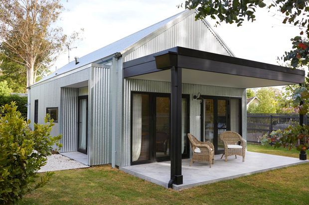 A compact contemporary pavilion-style house clad in pale corrugated metal, with a pitched gable roof, large glazed doors and windows, and a deep flat-roofed covered terrace supported by dark posts. Wicker chairs sit on the concrete patio, with a fenced garden and established planting surrounding the building.