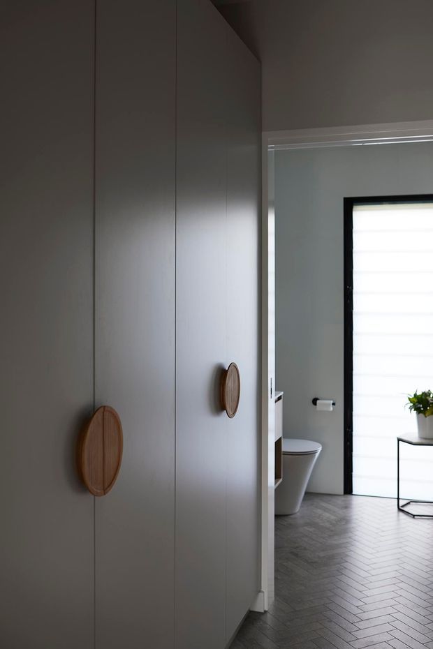 A moody contemporary bathroom viewed past a tall bank of flush grey cabinetry with distinctive round timber pulls. The bathroom includes a white toilet, grey wall surfaces, dark herringbone floor tiles, a black-framed window with a translucent roller blind, and a small black-framed side table with a potted plant.