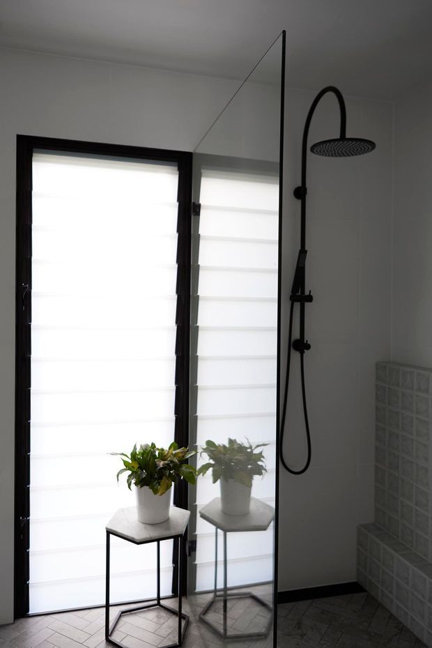 A contemporary bathroom with a frameless full-height glass shower screen, matte black rainfall shower and handheld fixture, dark-framed frosted window with white blinds, and a small potted plant on a slim black metal side table. The floor has dark patterned tiles, while a translucent glass-block tiled shower bench or wall feature is visible at right.