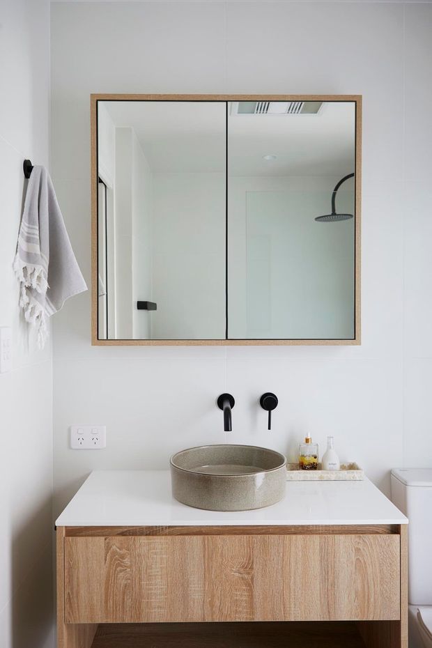 A bright, contemporary bathroom featuring a wall-mounted timber vanity with a white benchtop, round vessel basin, matte black wall-mounted tapware, and a large wood-framed mirrored cabinet. White wall finishes, a black-framed shower visible in the mirror, and a neutral textured towel complete the clean, restrained design.