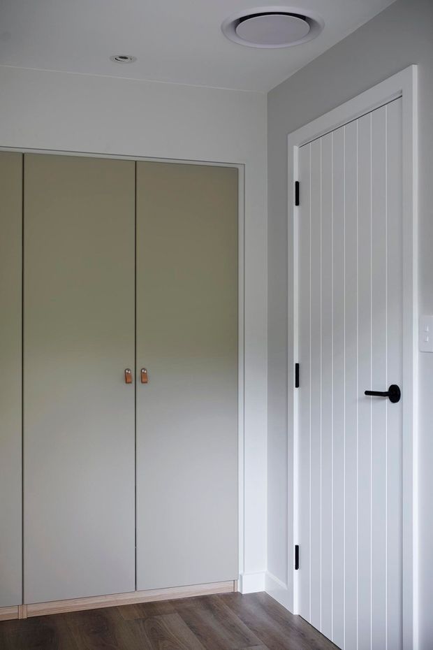 A clean, compact interior hallway or entry area with full-height built-in wardrobe cabinetry in a pale grey-beige finish, a white vertically grooved internal door with black hardware, white walls and trim, and dark timber-look flooring. A circular ceiling air vent is visible.