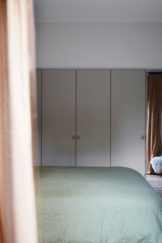 A softly lit bedroom viewed through a partially drawn curtain, featuring a neatly made bed with a muted pale green coverlet and a full wall of simple grey built-in wardrobe cabinetry. Brown curtains and a glimpse of a soft chair or bedding appear at the right edge, creating a calm, understated interior.