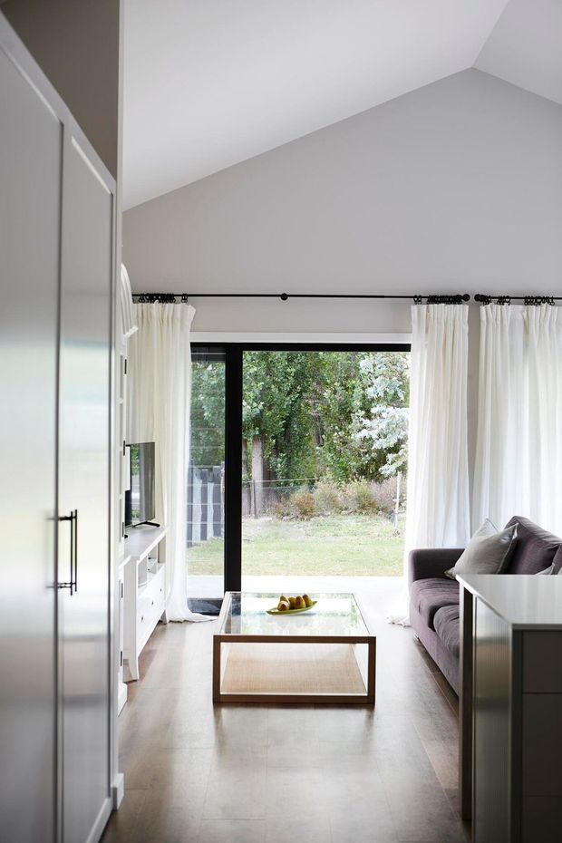 A bright, narrow contemporary living room with a high sloped ceiling, pale grey walls, light flooring, and full-height black-framed glazing opening to a garden. White sheer curtains frame the window, while a dark grey sofa, glass coffee table, white media cabinetry, and tall built-in storage line the room.