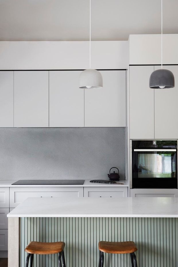 Contemporary kitchen with white flat-panel cabinetry, a grey patterned splashback, integrated black oven, white stone island benchtop, and a vertically fluted muted green island front. Two pendant lights and timber-topped stools add contrast.