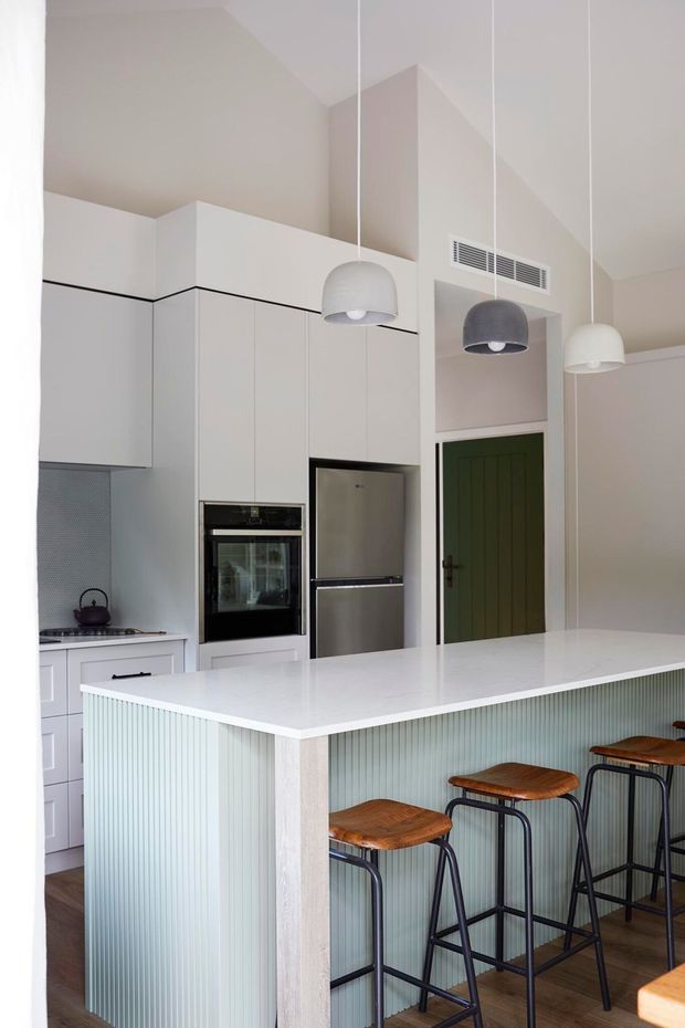 Bright contemporary kitchen with a large white island featuring a pale green vertically fluted base and seating for four on timber-topped black metal stools. The kitchen has streamlined white cabinetry, integrated stainless-steel appliances, a white benchtop, pendant lights, timber flooring, and a dark green panelled door.